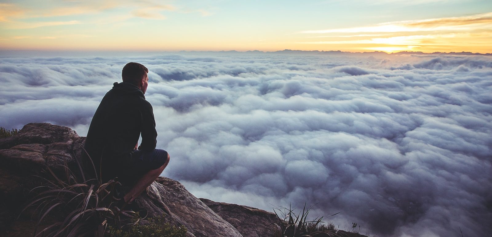man sitting on gray rock while staring at white clouds
